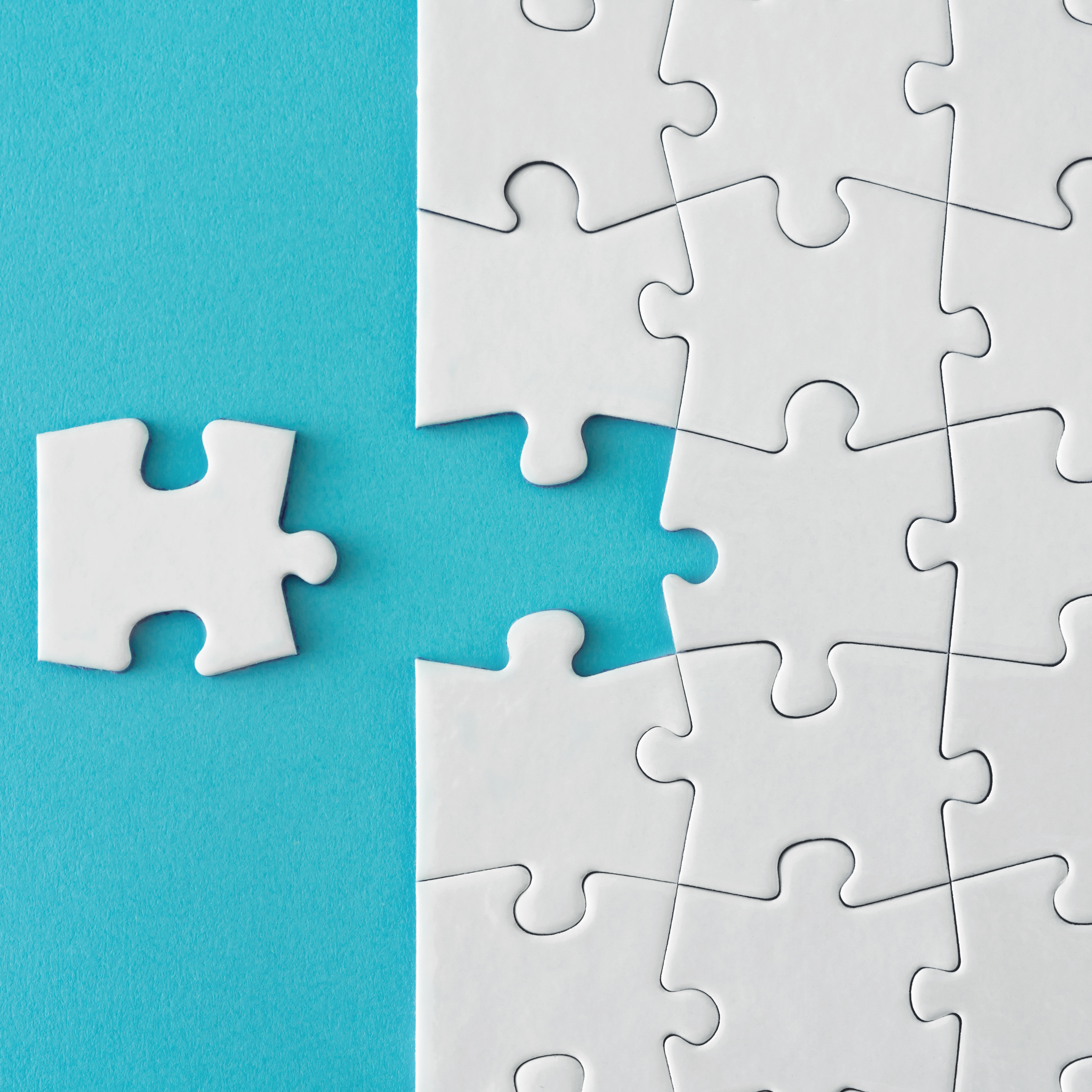 A single white puzzle piece sitting next to a nearly completed white jigsaw puzzle with one missing spot on a turquoise background.